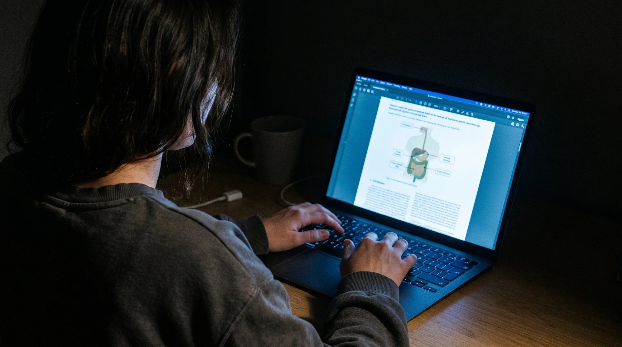 Laptop glowing in a dark room at night with a research paper on screen showing a diagram of the digestive system